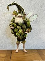 Full view of the Mark Roberts Fruit Fairy Doll standing up against a light wall, showing plush fruit-based costume, white beard, pointed hat, and fairy wings.
