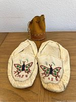 Pair of light-colored moccasins with detailed butterfly beadwork and the word 'TASK' on top, laid flat showing top view.