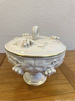 Front angle of white porcelain covered dish with sculpted grape clusters and gold trim on lid and base.