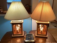 Photo showing both lamps with pleated beige lampshades and quartz clock with gold frame on a wooden table.