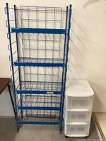 Photo showing the blue metal folding shelf next to the white plastic storage unit against a white wall.