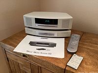 Front view of Bose Wave Music System III stacked with multi-CD changer, two remotes and a printed manual on top