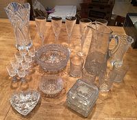 Overall view of assortment of cut and frosted glassware including pitcher, champagne glasses, vases, shot glasses, candy dish, and plates arranged on wooden table.