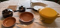Full view of all cookware items on table showing yellow Copco pot, brown pan, 2 Red Copper pans, 2 Utopia pots, and small black saucepan