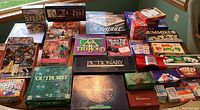 Photo showing the full lot including multiple board games, card games, and puzzles stacked on a table, highlighting titles like Scrabble, Pictionary, TriBond, Would You Rather?, and others.