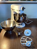 White Kenmore Elite stand mixer with a stainless steel bowl, power cable visible, placed on a wooden table along with an additional larger stainless steel bowl and three mixer attachments (wire whisk, dough hook, flat beater).