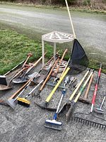 Full view of lot outdoors on pavement showing Rubbermaid tool stand and various garden tools including rakes, hoes, shovels, cultivators
