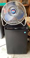 Wide shot showing the full Danby mini-fridge with closed door and Lakewood fan sitting on top.