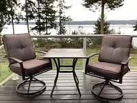 Patio set showing 2 swivel chairs and square table on an outdoor deck with a lake and trees in the background.