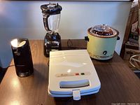 Lot of four small kitchen appliances arranged on table: Black & Decker can opener, Hamilton Beach blender, Toastmaster Belgian waffle maker closed, Rival crockpot