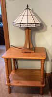 Photo showing the small wooden table with turned legs, rectangular top, and bottom shelf. The stained glass style lamp sits on top, cord visible.