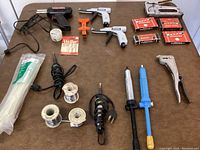 Photo of soldering tools, solder wire reels, staple guns, staples, long cable ties in packaging, and other fastening tools arranged on brown surface.