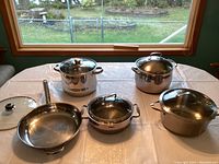 Five stainless steel pots and pans visible on a table, showcasing the full lot contents including the stock pots, steamer, sauté pan, and covered casserole dish with glass lids.