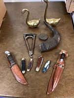 Overview of the lot including all knives, brass geese, horn, medallion, and multi-tool on a brown surface.