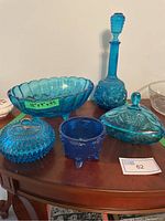 Photo showing entire set of five blue glass serving pieces on wooden surface, including decanter with stopper, oval serving bowl with label showing dimensions, two lidded candy dishes, and small open dish.