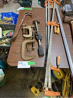 Overall view of assortment of clamps on brown table: includes bar clamps, C-clamps, and claw clamps with varying wear and rust.