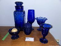 Full assortment of cobalt blue glass pieces including vases, bowls, lidded jar, and small cups arranged on wooden surface.