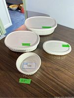 Four CorningWare French White stoneware pieces on wooden surface. Three with rubber lids, one bowl without lid. Fragile condition noted.