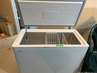 White Danby chest freezer with open lid showing the interior and one removable wire basket inside, photographed in a basement area.