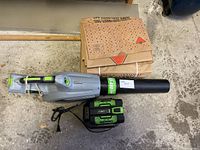 Full view of the 56V EGO Power+ blower with attached tube, battery and charger in front, and a stack of folded brown paper yard waste bags behind on concrete floor.