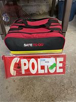Closed red and black Safe To Go emergency kit bag sitting on floor next to Goodwrench Police sign.