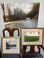 Group photo of three framed artworks displayed on wooden chairs: large canvas print of snow-covered road, a green field painting, and a forest-lake scene.