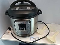 Instant Pot 6 quart electric pressure cooker with power cord, two white plastic spoons, and clear measuring cup on white shelf.