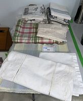 Stack of various folded kitchen linens including white embroidered tablecloths, plaid placemats, and lace-edged cloths
