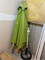 Full view of folded green patio umbrella with white metal pole leaning against wall.