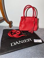 Red leather handbag with dual rounded handles and tassel, displayed on a black Danier storage bag alongside an adjustable long strap with metal clasps.