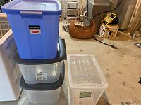 Four storage bins stacked on top of each other. Two blue large bins and two clear bins with grey lids visible on concrete floor.