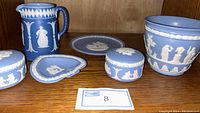 Full set of blue jasperware pieces arranged on wooden shelf, showing pitcher, two lidded boxes, plate, heart-shaped dish, and bowl with white reliefs.