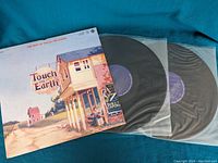 Photo of the album cover and two black vinyl records in clear protective sleeves lying on a blue fabric background.