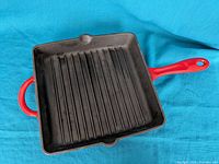 Top view of square cast iron grill pan with red enamel handle and red exterior sides. Black interior with parallel raised ridges and pouring spouts.