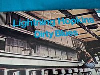 Front cover of the Lightning Hopkins Dirty Blues vinyl record. The image shows Lightning Hopkins sitting next to a large truck in an industrial environment. The title and artist name are printed in bright blue.