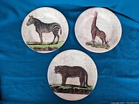Full view of all three Kenya series plates laid out on blue fabric showing zebra, giraffe, and leopard designs.