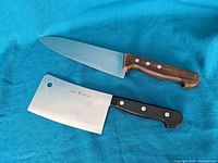 Two J.A. Henckels kitchen knives displayed on blue fabric, showing one cleaver with a black handle and a chef's knife with a wooden handle.