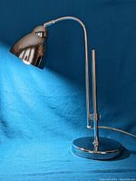 Full view of chrome modernist table lamp with base, adjustable rods and curved arm with cone-shaped shade