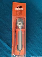 Photo of JVC MD-247 microphone in sealed plastic blister pack with orange card backing, showing the microphone's silver body and mesh grille.