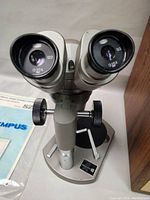 Front view of microscope showing dual eyepieces labeled Ø20X on adjustable zoom head tube atop the vertical column and black stage plate.