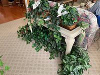 Overview of six artificial plants in baskets with trailing vines and broad leaves, some with birds and red berries, on sunroom furniture and floor.