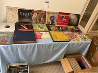 Photo showing multiple vinyl records laid out on a table and a box with cassette tapes underneath, some record album covers visible including holiday music and classic vocal groups.