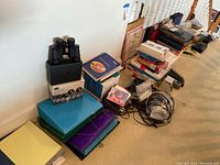 Full view of the lot showing binoculars on top of case and box, stacks of various colored accordion file folders, cables, books, and office supplies.