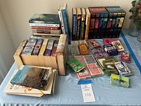 Tablescape showing two boxed 'Left Behind' book sets, multiple James Patterson books, several Catholic-themed books, and a large assortment of vintage playing cards.