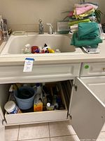Wide view of laundry sink with various cleaning supplies inside the sink and a stack of colorful cleaning rags on the counter.