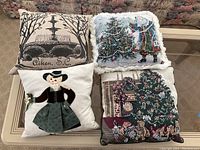 Four decorative square pillows on a table; includes a pillow with fountain in Aiken, SC, a historical woman appliqué, and two Christmas-themed pillows with embroidery and fringe.