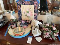 Full view of the lot showing various decorative tins, plush lamb, vanity tray, floral ring and more on a wooden table.