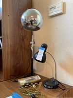 Photo shows the smaller black flexible neck lamp labeled '121' next to cords and a box containing spare parts or bulbs, set on a wood surface near a wall thermostat.