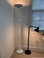 Photo showing white torchiere floor lamp with round base and a black adjustable reading lamp next to window with blinds