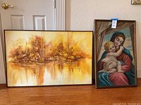 Photo of two framed paintings placed on the floor against a wall. Left painting is a village scene with houses, trees, and reflective water in sepia yellow tones. Right painting is a Madonna holding Child Jesus with warm colors in a frame.
